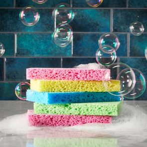 Colorful sponges stacked, covered in soap foam, surrounded by floating bubbles on a marble counter with a blue tiled wall backdrop.