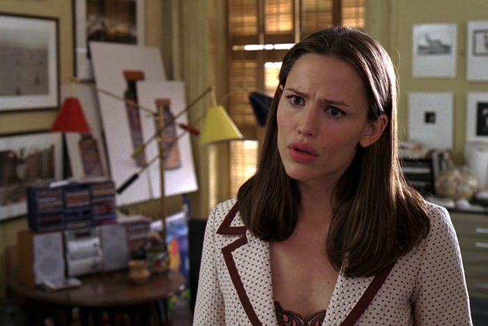 Woman standing confused, in an art-filled room with framed pictures, sketches, and CDs on a wooden table in the background.