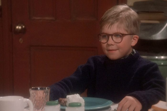 A young boy wearing glasses smiles while seated at a dining table, surrounded by dishes in a cozy room with a wooden door.