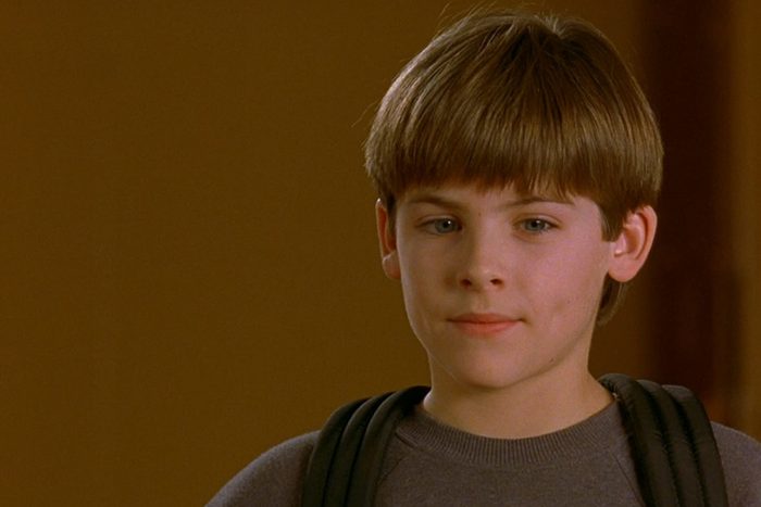 A young boy with short hair stands calmly, wearing a gray sweater, against a plain brown background.