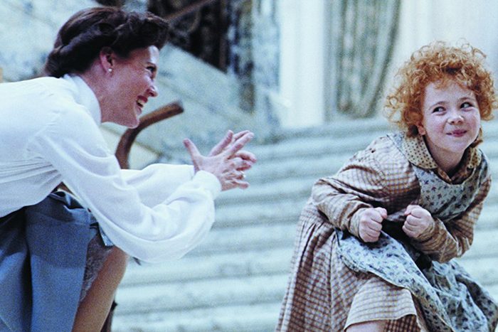 A woman smiles and claps at a curly-haired child posing playfully on stairs in a bright environment.