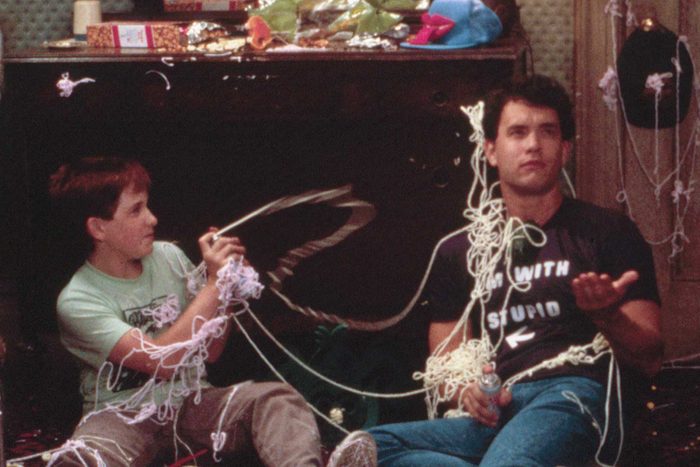Two people sit tangled in colorful streamers, playfully spraying them. The room is messy with scattered boxes and items. One shirt reads 