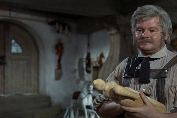 A man holds a wooden puppet in a rustic room with wooden beams, a door, and a rocking horse visible in the background.