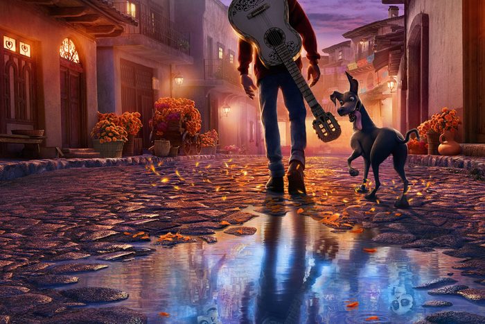 A boy with a guitar walks a dog down a cobblestone street, surrounded by flowers and warm lantern light reflecting in a puddle.