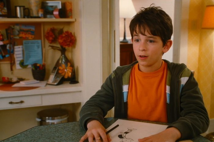 A young person sits at a table drawing, with a cluttered desk and telephone in the background.
