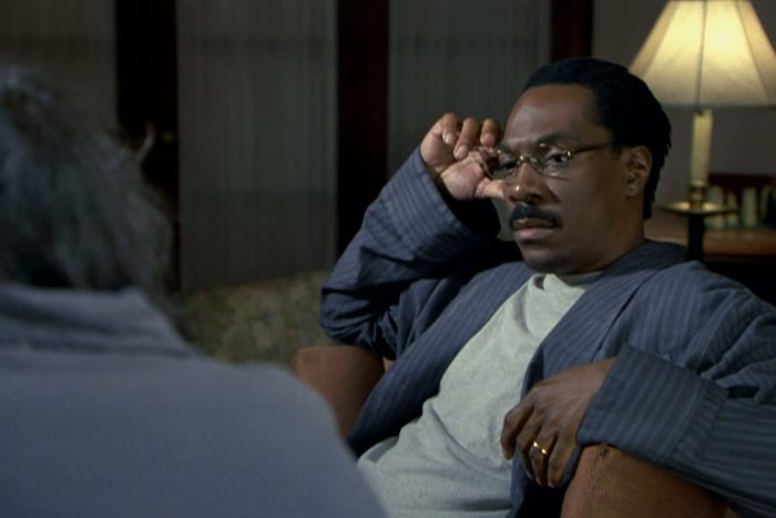Man adjusts glasses, seated attentively, in a cozy, dimly-lit room with a lit table lamp in the background.