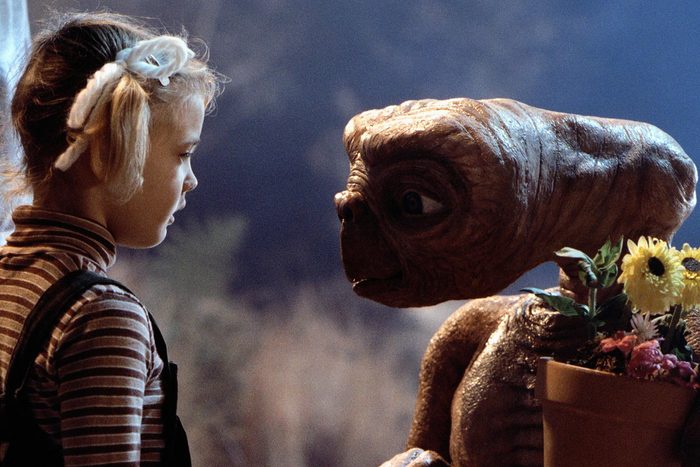 A creature holds a flower pot, interacting with a young girl in striped clothing, set against a blurred outdoor background.