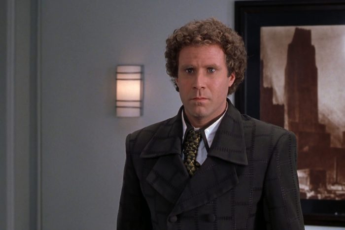 Man stands expressionless indoors, wearing a patterned suit and tie, with a framed cityscape artwork and wall sconce in the background.