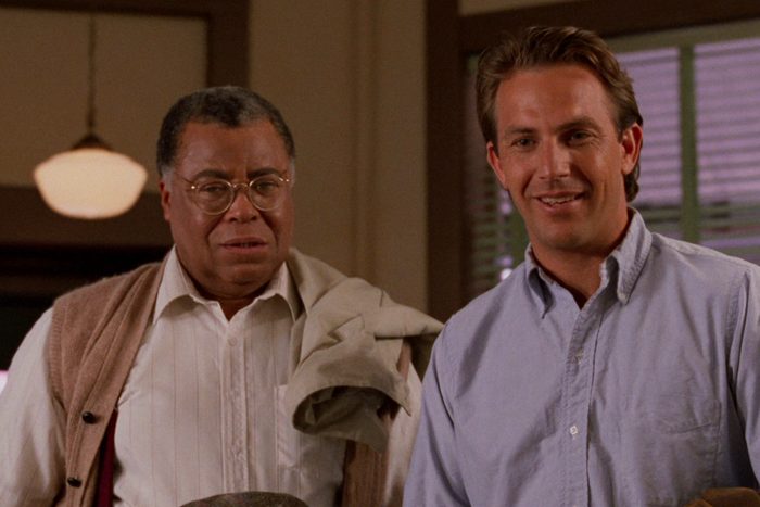 Two men stand together, one holding a coat, indoors with soft lighting and wooden window blinds in the background.
