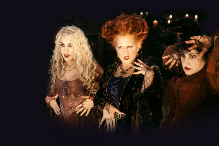 Three women with dramatic makeup and costumes stand indoors, striking theatrical poses with expressive faces, surrounded by dim candlelight.