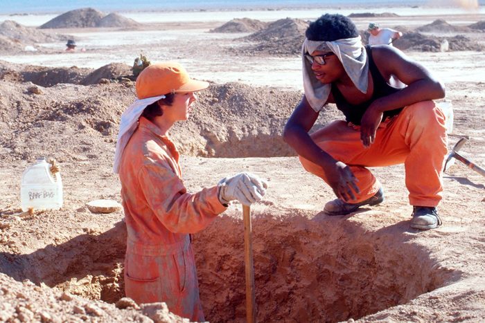 Two people in work clothes rest near a dug hole in a dusty, barren landscape. A nearby jug reads 