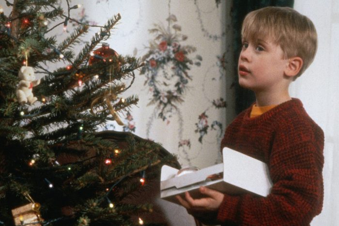 Child holding ornament stands by decorated Christmas tree with lights and teddy bear ornament, in room with floral wallpaper.