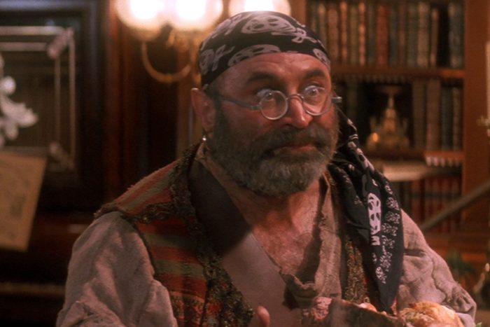 Bearded man wears glasses, pirate attire, and a skull bandana, in a warmly lit room with bookshelves and lamps.