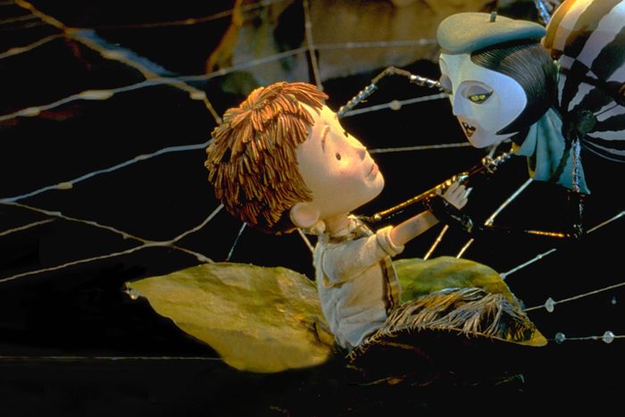 Boy interacts with a human-faced spider on a giant leaf, surrounded by a web in a dimly lit setting.