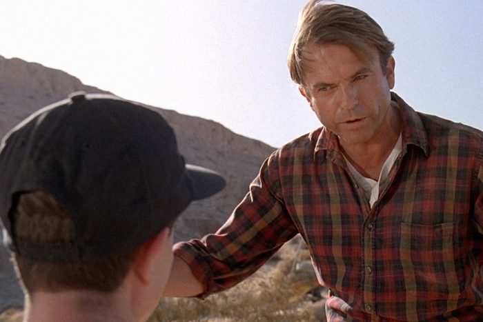 A man in a plaid shirt gestures while talking to a person in a cap in a rocky outdoor setting.