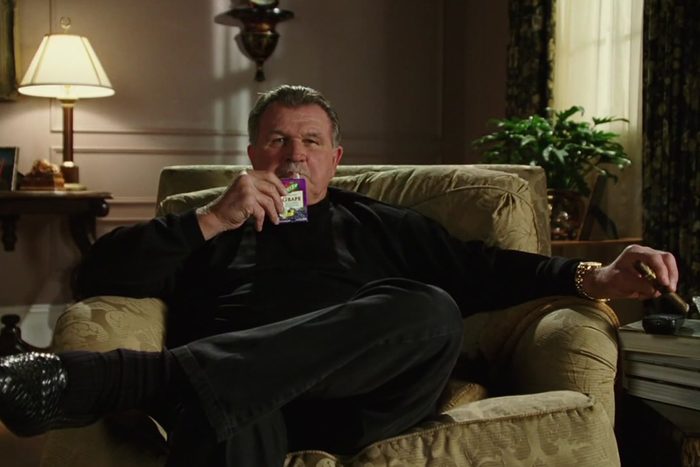 Man relaxing, holding grape-flavored snack, seated on upholstered armchair. Room features a lamp, side table, plant, and patterned curtains.