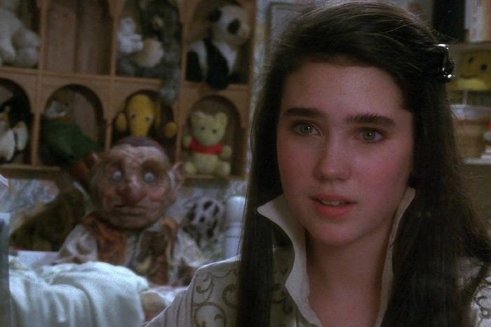 Young woman looks forward; a puppet-like creature sits behind her; surrounded by various stuffed animals on shelves.