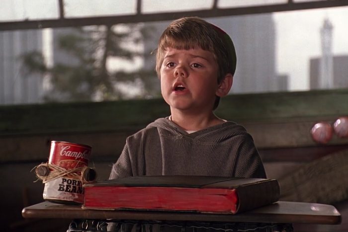 Child sits at a classroom desk, speaking, with a Campbell's Pork & Beans can and a closed book. Blurred windows in the background.