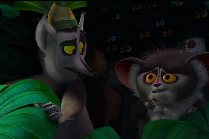 Two animated lemurs, one wearing a hat, look concerned amidst green foliage with glowing eyes peering from the darkness.