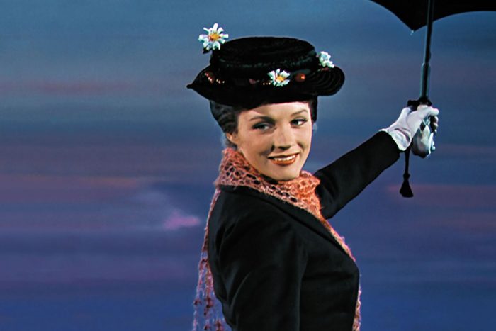 A person holds an umbrella, wearing a black hat with flowers, smiling, against a cloudy sky backdrop.