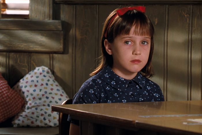 Girl sits quietly, wearing a red headband and blue floral dress, in front of a wooden wall and cushioned seating.