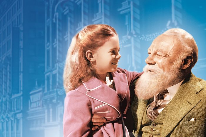 A child smiles at an older man with a beard, holding her. They're dressed formally with a blue background depicting a cityscape.