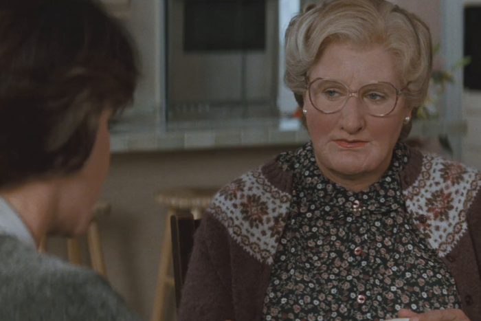 Person wearing glasses looks attentively, sitting in a kitchen with floral-patterned sweater and shirt. Another individual is partially visible at the table.