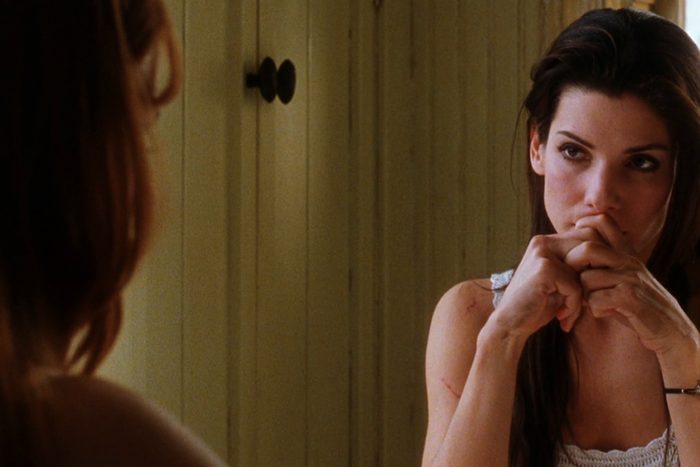 Person sits pensively, hands clasped near face, in a room with wooden paneled walls, partially mirrored reflection visible.