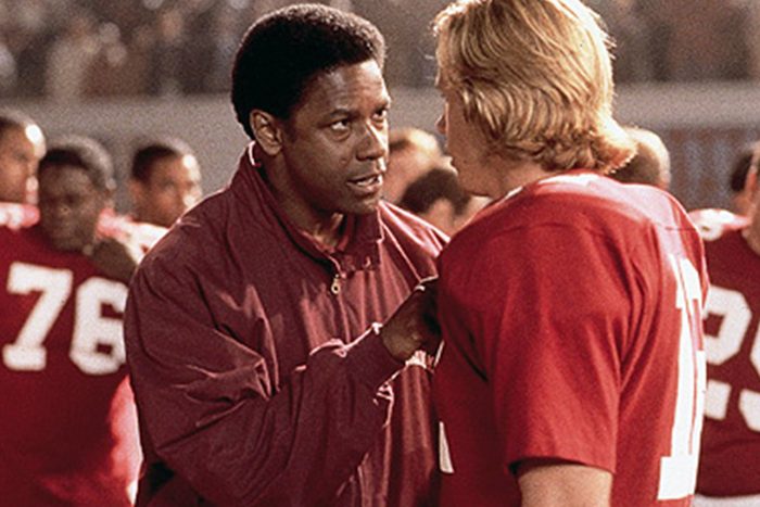 A man in a maroon jacket talks intently to a football player wearing a red jersey amidst a crowded stadium.