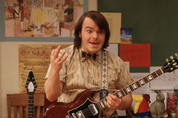A person plays the guitar animatedly, in a classroom adorned with colorful papers and wall posters.