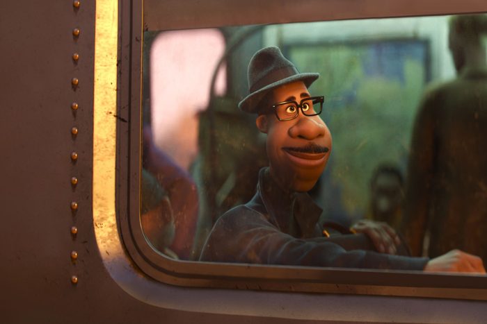 Animated character wearing glasses and a hat, smiling while looking out of a train window, with blurry passengers in the background.