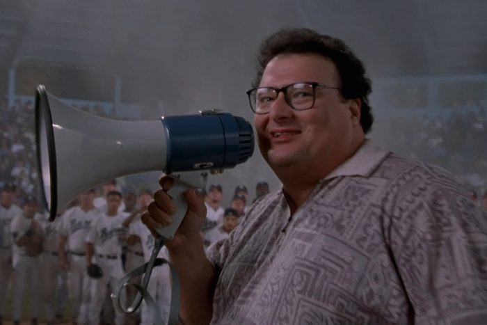 Person holding a megaphone is smiling, standing in a baseball stadium filled with players and spectators in the background.