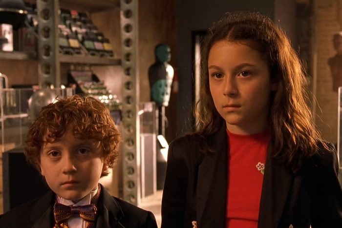Two children stand quietly in a room filled with shelves of gadgets and technology, giving a focused and serious expression.