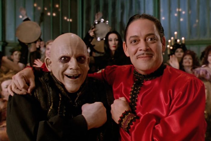Two smiling men, one bald and the other with a mustache, stand arm-in-arm. They are in a lively setting with people and warm lighting.