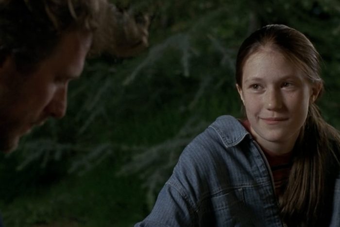 Two people appear outdoors; one looks at the other, who is smiling slightly. They're surrounded by greenery, suggesting a natural setting.