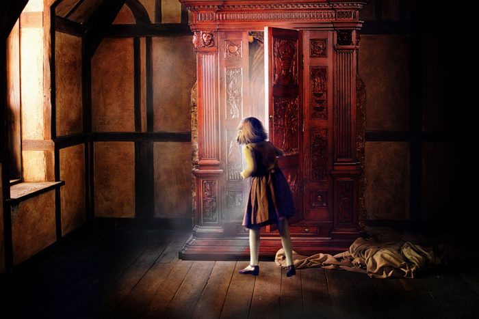 A girl opens an ornate wardrobe, light streaming out, in a dimly lit, wooden-paneled room with a draped fabric on the floor.