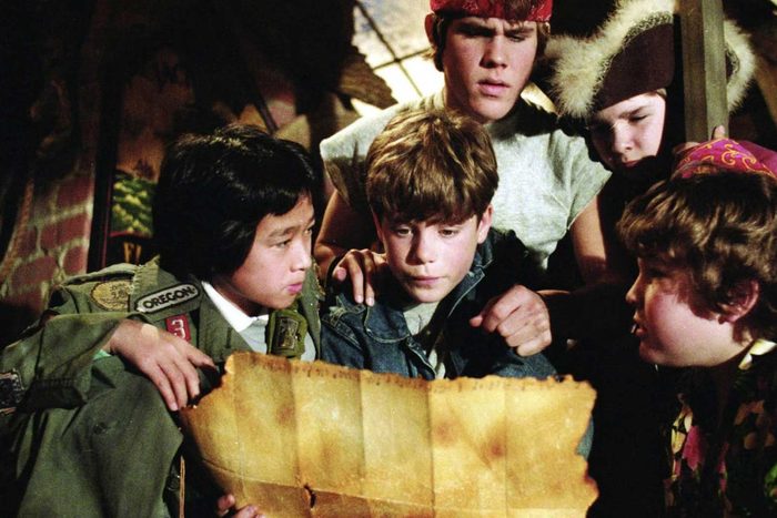 Five kids examine a worn map intently, huddled together in a dimly lit attic with brick walls and ropes.