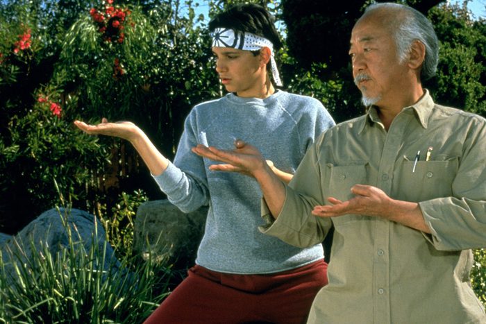 Two people perform a balancing exercise in a garden, each with one arm extended, focusing intently on their open palms.