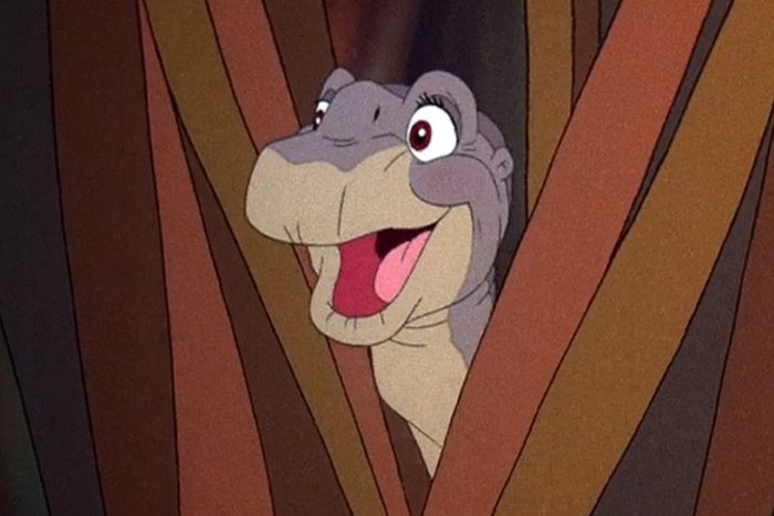 Animated dinosaur smiles widely, poking head out between tree trunks in a forest setting.