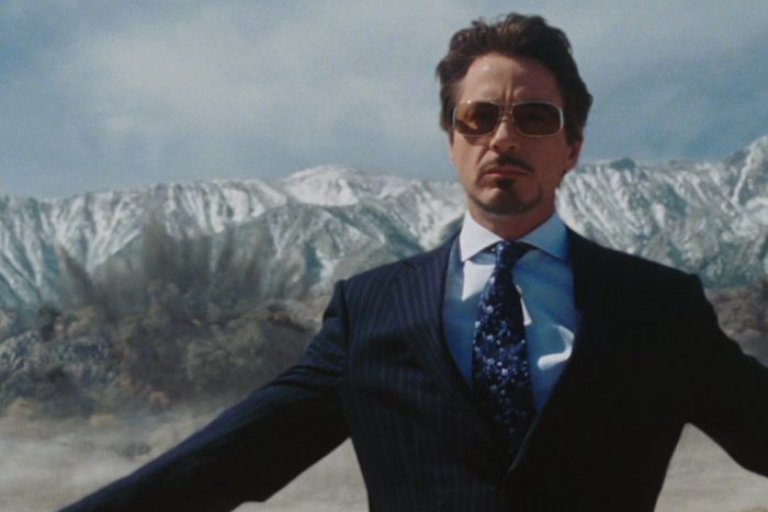 Man in suit stands confidently, arms outstretched, before snowy mountains while a large explosion occurs behind him.