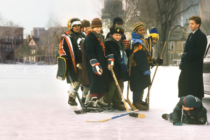 A group of children, dressed in winter clothing, stand on an ice rink with hockey sticks, while one child sits on the ground nearby. Cityscape visible.