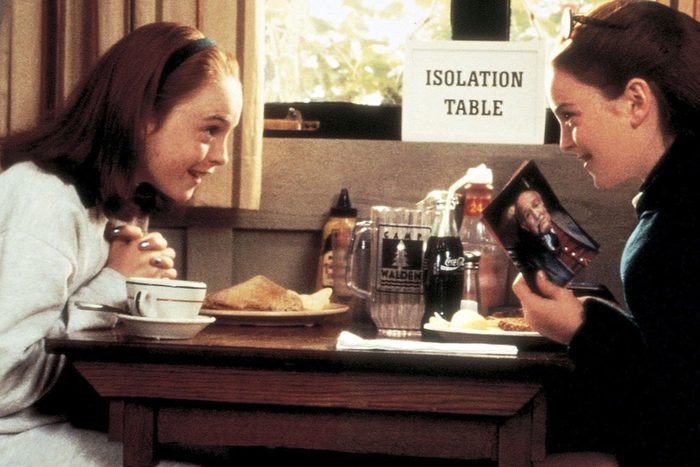 Two girls talk at a table with food, one holding a photo. A sign reads 