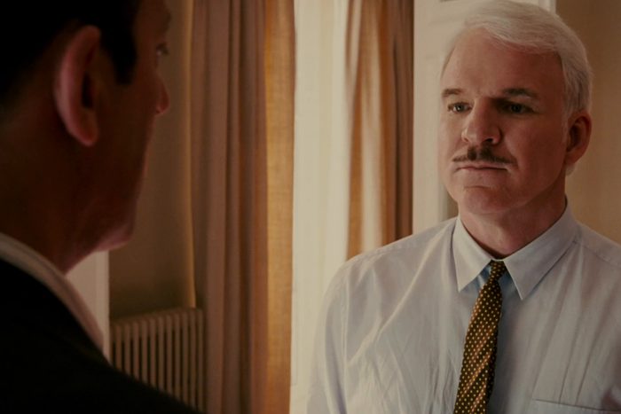 A man with white hair and a mustache looks serious in a room with beige curtains, wearing a shirt and tie.