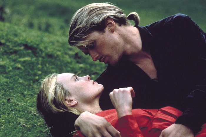 Two people lie on grassy ground, gazing at each other closely. One wears a red garment, while the other wears black.