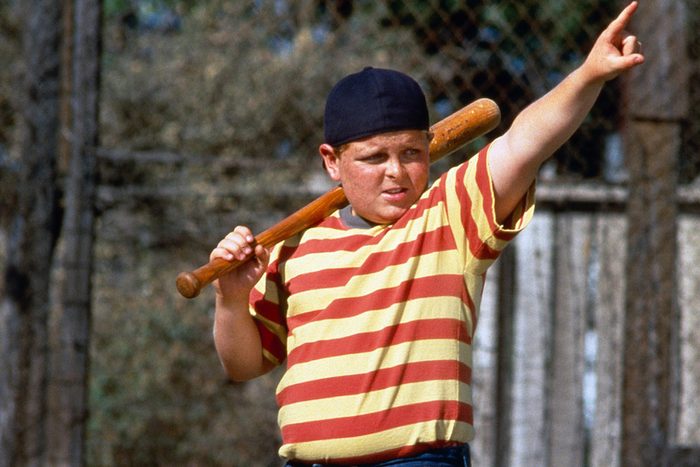 A person holds a baseball bat on their shoulder, pointing forward, wearing a striped shirt and hat, outdoors by a chain-link fence.