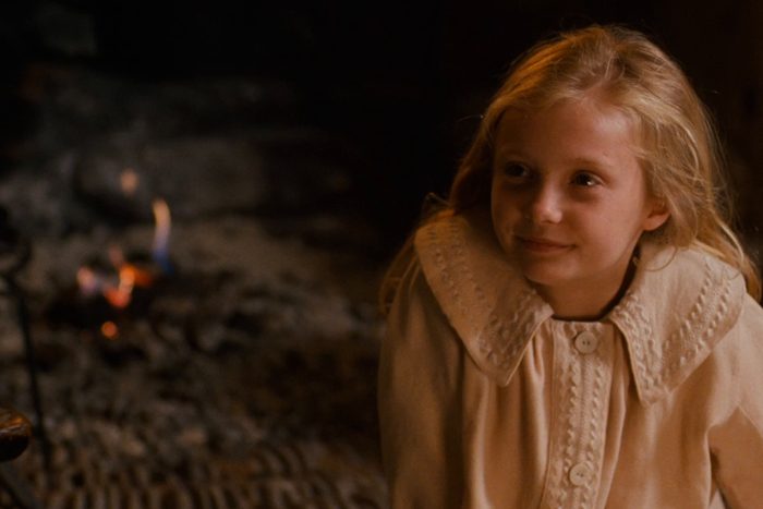 Girl wearing a cream-colored, embroidered coat sits smiling, with a fireplace glowing softly in the dim background.
