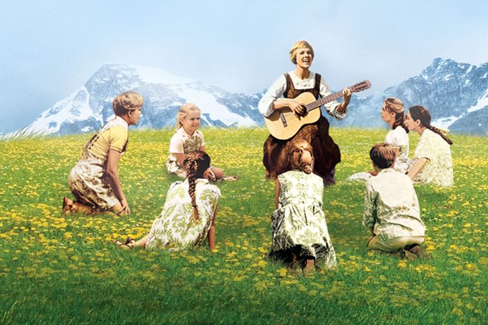 A person plays guitar while children sit in a circle on a flower-covered field, with snowy mountains in the background.