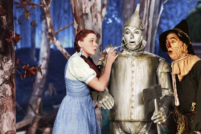 A girl oils a tin man in a forest, while a scarecrow looks on with surprise.