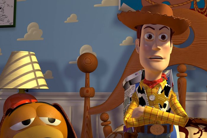 Cowboy toy stands with arms crossed, looking serious. Background features a lamp and cloud-themed wallpaper near a bed.