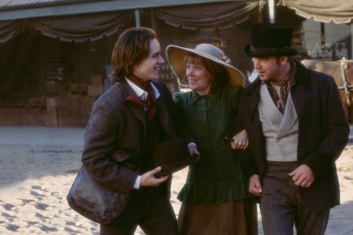 Three people, dressed in period clothing, smile and walk arm-in-arm on a sandy street with a market stall in the background.